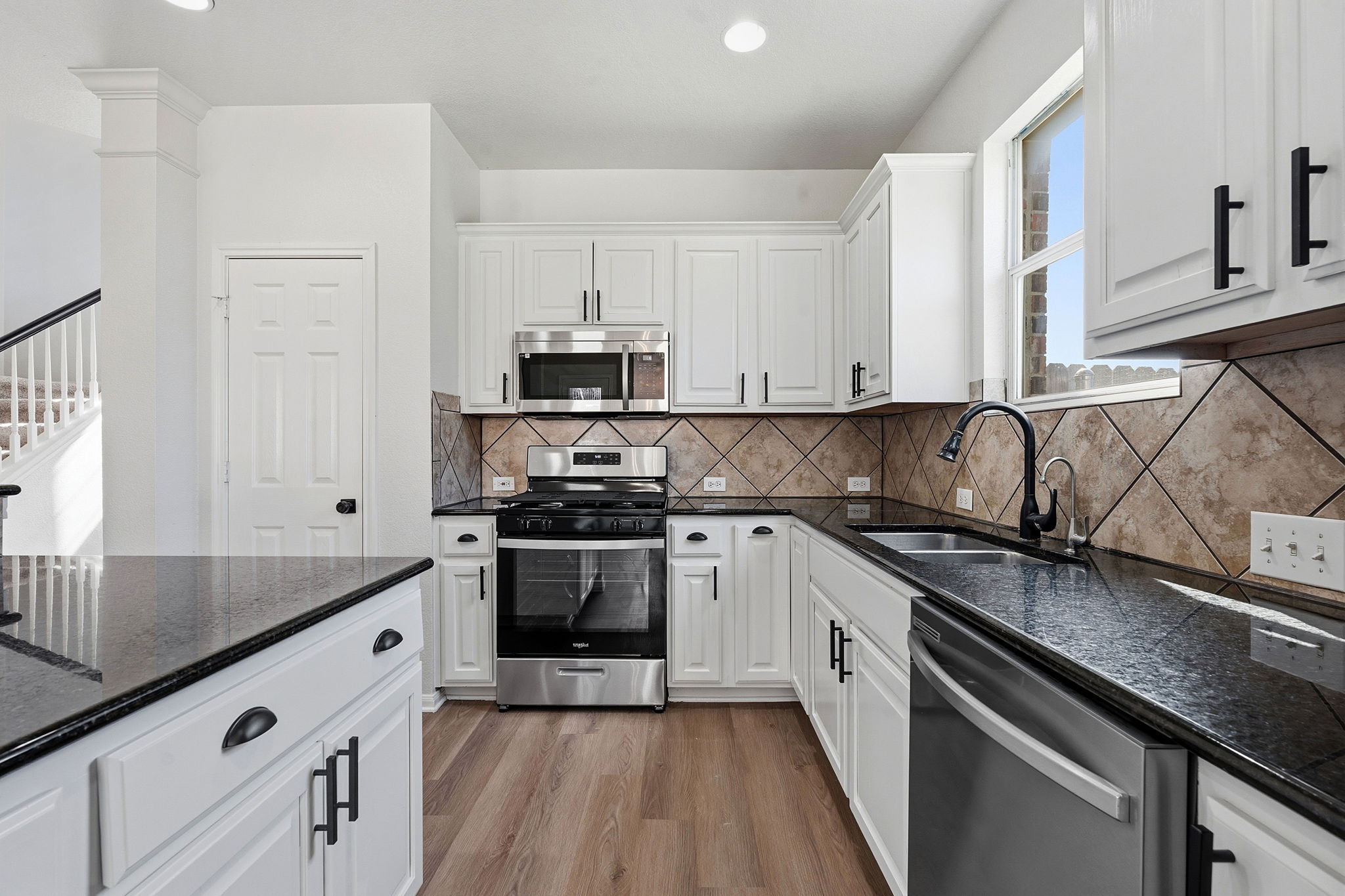 Bright and inviting kitchen featuring white shaker cabinets, dark stone countertops, stainless steel appliances, and a stylish diagonal tile backsplash. Natural light from the window and warm wood-look flooring create a clean, modern space that's both functional and welcoming.